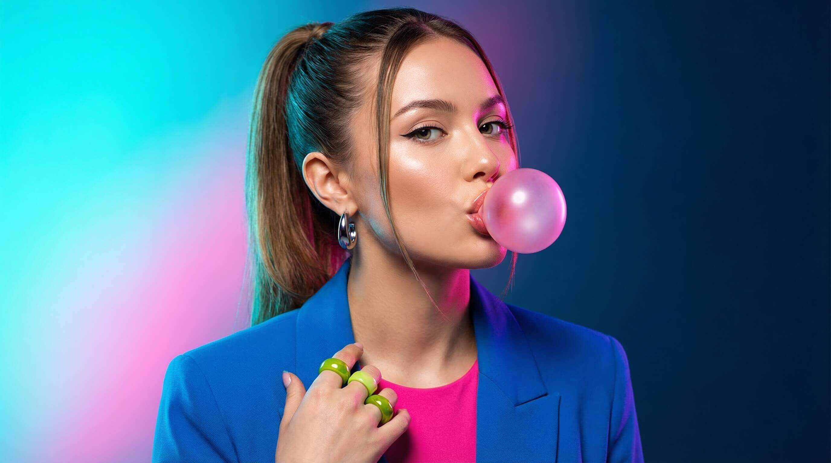Стильная девушка в ярком образе надувает розовый пузырь жвачки